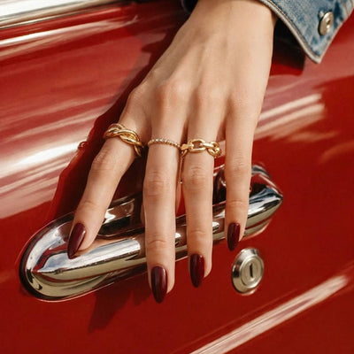 A close-up of a hand with dark nail polish and gold rings resting on the chrome door handle of a red vintage car, wearing a blue denim sleeve.
